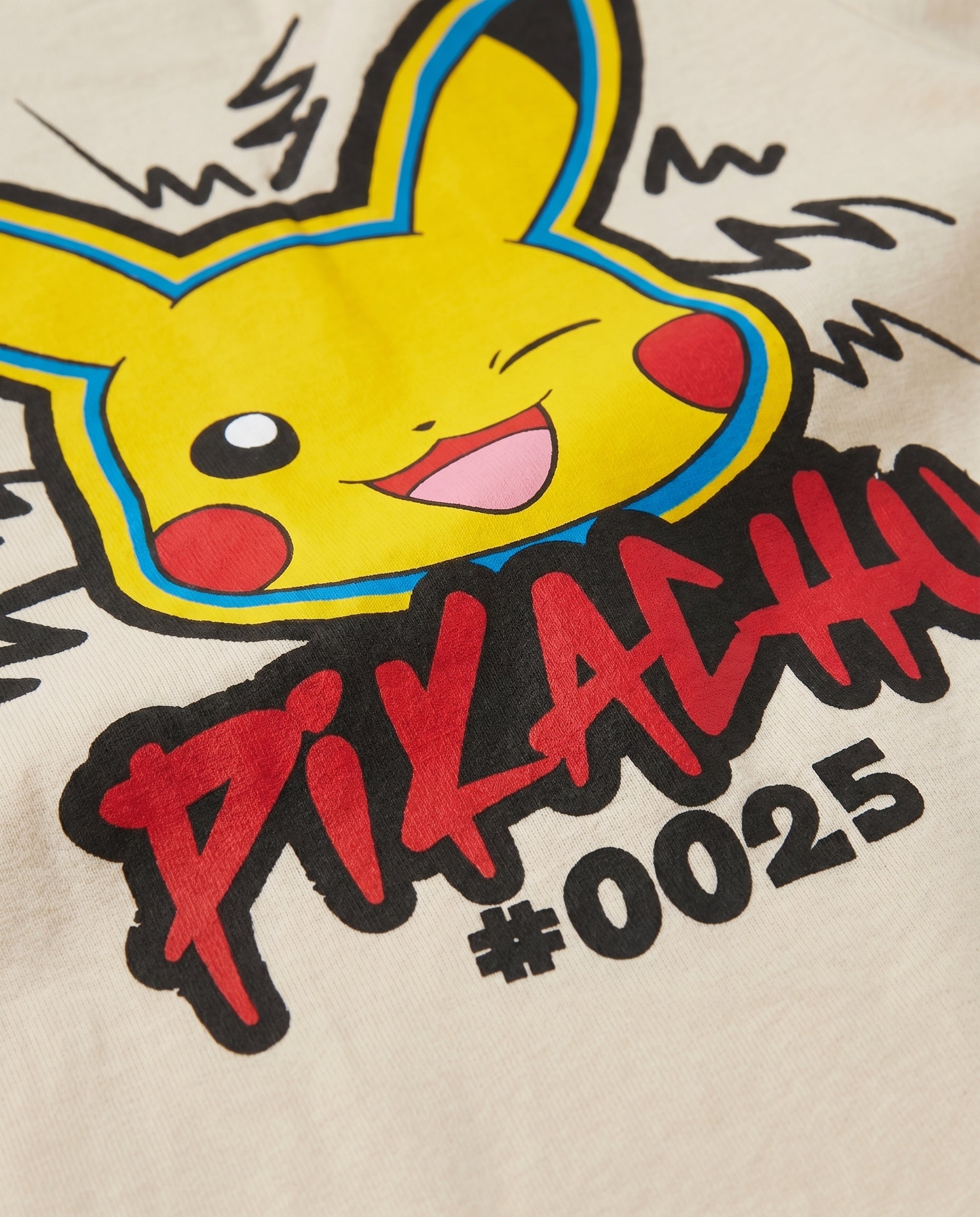 Close-up of the back print on a cream kids' t-shirt showing a winking Pikachu character face with red graffiti lettering and #0025 text