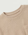 Close-up neckline and texture detail:
Close-up of the ribbed crew neckline and drop shoulder construction on a kids' sand waffle cotton t-shirt, highlighting the fine raised grid texture of the waffle knit fabric and tonal tack stitching detail.