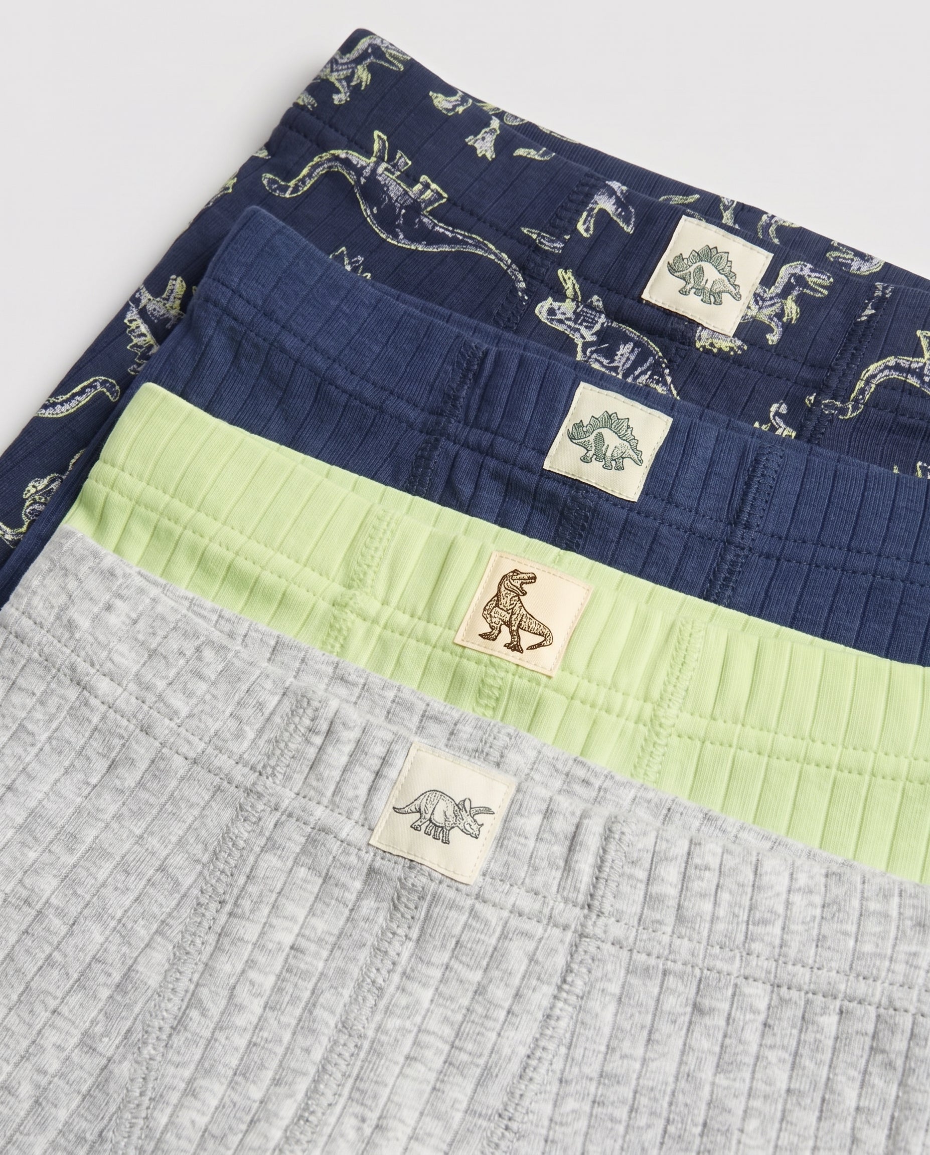 Stacked waistbands of boys' ribbed boxer briefs in navy dino print, solid navy, lime green, and grey marl showing dinosaur woven labels