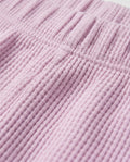 Close-up of a Toddler girls' waffle-knit cotton short pants in pink lilac with a wide elasticated waistband, laid flat on a light grey background.