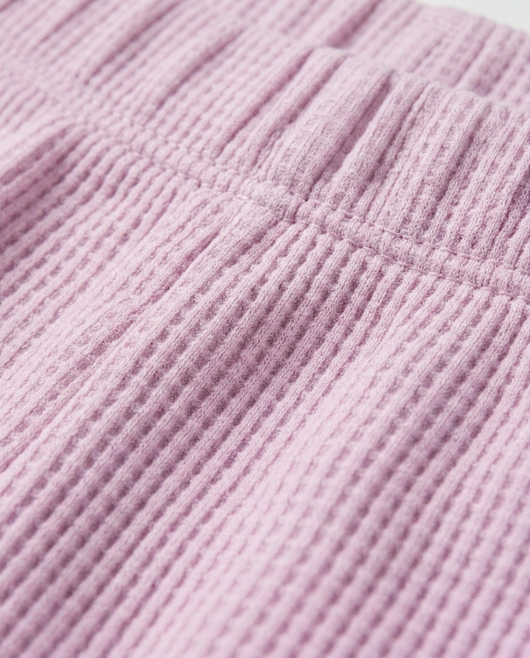 Close-up of a Toddler girls' waffle-knit cotton short pants in pink lilac with a wide elasticated waistband, laid flat on a light grey background.