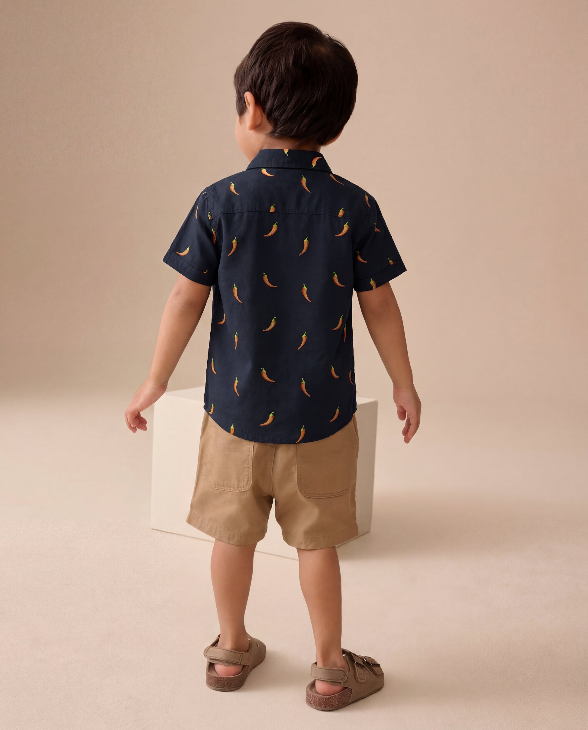 Child wearing a navy shirt with chilli pattern and beige shorts on a beige background